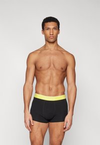 Svarta boxertrosor med ett gult midjeband som har "LACOSTE"-logotypen; slät textur, tät passform och med längd till mitten av låret.