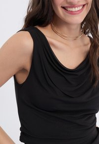 Sort ærmeløs top med draperet udskæring, glat tekstur, tætsiddende design og minimal syning. Guld lagdelt choker halskæde.