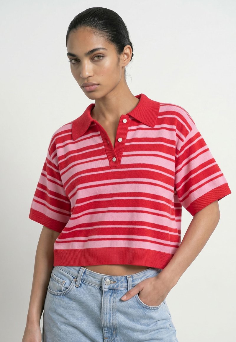 Femme aux cheveux foncés portant un polo rayé rouge et rose à manches courtes et un jean bleu clair, une main dans la poche.