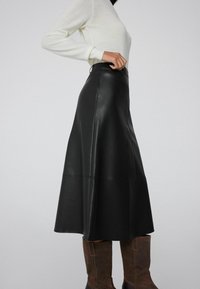 Falda midi de cuero negro con un ligero vuelo, que presenta una textura suave y una cintura ajustada. Combinada con botas marrones hasta la rodilla.