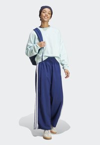Himmelblaues Baumwoll-Sweatshirt, marineblaue weit geschnittene Hose mit weißen Seitenstreifen, weiße Sneaker und eine marineblaue Mütze. Modell trägt eine blaue Tasche.