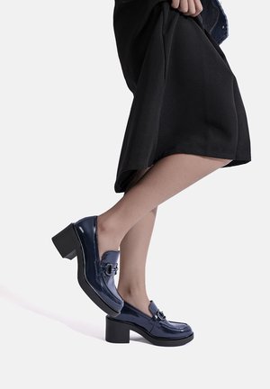 Persoon draagt glanzende marineblauwe loafers met hak en een zwarte rok, tilt één voet iets van de grond tegen een witte achtergrond.