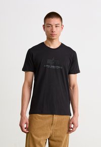 Svart bomull T-skjorte med rund hals, med en strukturert grafisk utskrift av "ALPHA INDUSTRIES" på forsiden. Modell iført beige fløyelsbukser.