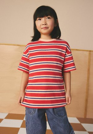 Ung flicka med rakt svart hår som bär en röd, blå och vit randig t-shirt och blå jeans, stående på ett schackrutigt golv.