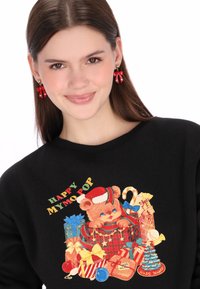 Fekete pulóver, medve grafikával, színes ajándékokkal, szalagokkal és ünnepi elemekkel körülvéve. A modell piros masni formájú fülbevalót visel.