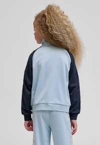 Laps pikkade lokkis juustega, seljas helesinine ja must tracksuit, seisab seljaga vaataja poole ühtlase tausta ees.