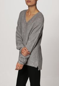 Harmaa neulepaita, jossa on V-pääntie, pitkät hihat ja väljä istuvuus. Siinä on hienovaraisia kimalle yksityiskohtia ja lantiolle ulottuva muotoilu.