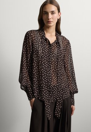 Jonge vrouw die een doorschijnende zwarte blouse met witte stippen en lange mouwen draagt, gecombineerd met losse zwarte broek, staand tegen een effen achtergrond.