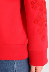 Sudadera roja con bordado floral en un patrón texturizado. Puños y dobladillo acanalados, con una etiqueta de marca en el lateral. El material parece suave.