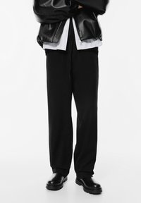 Persoon in zwart leren jack, witte shirt, losse zwarte broek en zwarte laarzen die met gekruiste armen tegen een witte achtergrond staat.