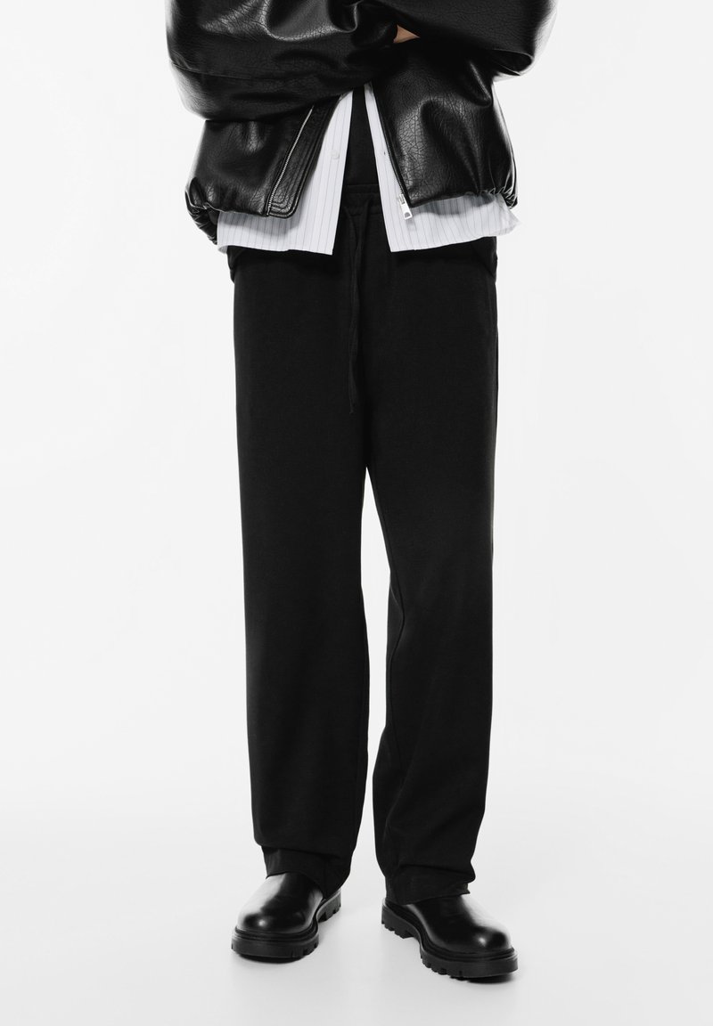 Persoon in zwart leren jack, witte shirt, losse zwarte broek en zwarte laarzen die met gekruiste armen tegen een witte achtergrond staat.