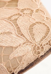 Gros plan d'un tissu en dentelle beige avec des motifs floraux et un fin filet sur un fond clair uni.