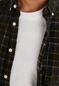 Grün-braunes kariertes Hemd zum Knöpfen, getragen über einem weißen T-Shirt. Das Hemd hat Knöpfe, einen Kragen und eine Tasche auf der linken Seite.