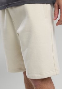 Lyhyt beige puuvillahousut pehmeällä tekstuurilla, joissa on sivutaskut ja suora leikkaus. Helma on ommeltu kestäväksi.