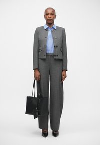 Blazer gris ajusté avec de grands boutons, chemise rayée bleue, pantalon gris à jambe large et sac fourre-tout noir. Des chaussures noires à bout pointu complètent la tenue.