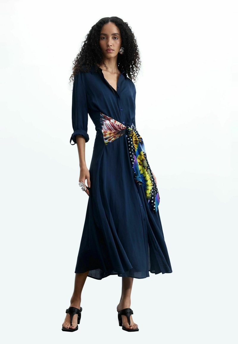 Femme aux cheveux bouclés portant une robe midi bleu marine avec des manches retroussées, une ceinture foulard colorée et des sandales noires à talons, debout devant un fond blanc.