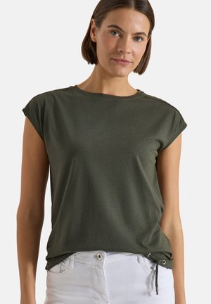 Femme aux cheveux courts bruns portant un haut vert foncé à manches courtes et un pantalon blanc sur un fond clair uni.