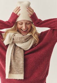 Beige stickad mössa och halsduk med en övergång från cream till ljusbrun, bärs över en lös röd tröja av mjukt, strukturerat tyg.