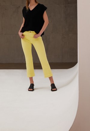 Frau trägt ein schwarzes ärmelloses V-Ausschnitt-Top, knallgelbe kurze Jeans und schwarze Sandalen mit offenen Zehen, steht vor einem schlichten Hintergrund.