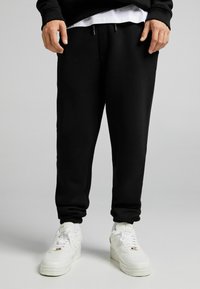 Black cotton blend sweatpants with elastic cuffs and drawstring waist, paired with white sneakers featuring a textured sole and logo accents.