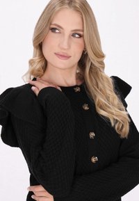 Zwarte gebreide cardigan met een gestructureerd patroon, gerimpelde schouders en vier ronde bruine knopen aan de voorkant. Lange mouwen.