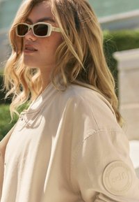 Donna bionda con occhiali da sole beige e un giubbotto beige con una toppa rotonda sul maniche, all'aperto alla luce naturale.