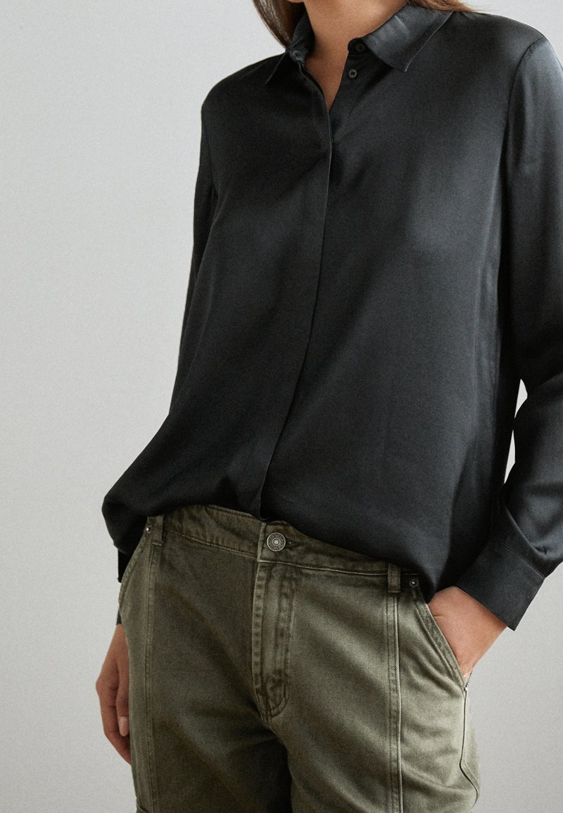 Chemise noire à manches longues, boutonnée, en tissu lisse, avec un col classique et une coupe décontractée, associée à un pantalon vert olive.