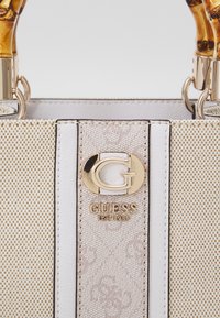 Borsa a tracolla beige con motivo, texture liscia, hardware dorato e manici in stile bambù. Presenta una targhetta con il logo "GUESS EST 1981."