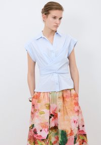 Vaaleansininen napillinen paita, jossa on hihatommat hihat ja kääntömuotoilu, yhdistettynä kukkakuosisiiin hameeseen, jossa on vaaleanpunaisia ja vihreitä sävyjä.