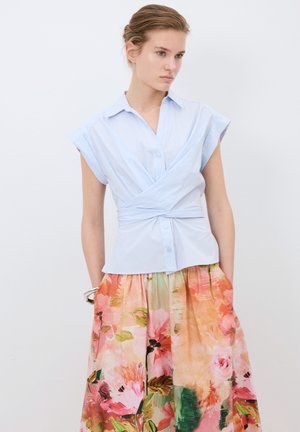 Camicia azzurro chiaro, con bottoni e maniche a cappuccio, abbinata a una gonna con stampa floreale nei toni del rosa e del verde.