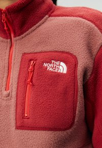 Gros plan d'une veste polaire rouge et rose avec une poche poitrine zippée et le logo The North Face brodé en blanc.