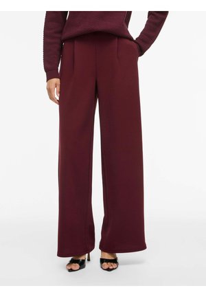 Kobieta w bordowych spodniach z szerokimi nogawkami, dopasowanym swetrze z dzianiny oraz czarnych, otwartych na palce szpilkach, stojąca z ręką na biodrze.