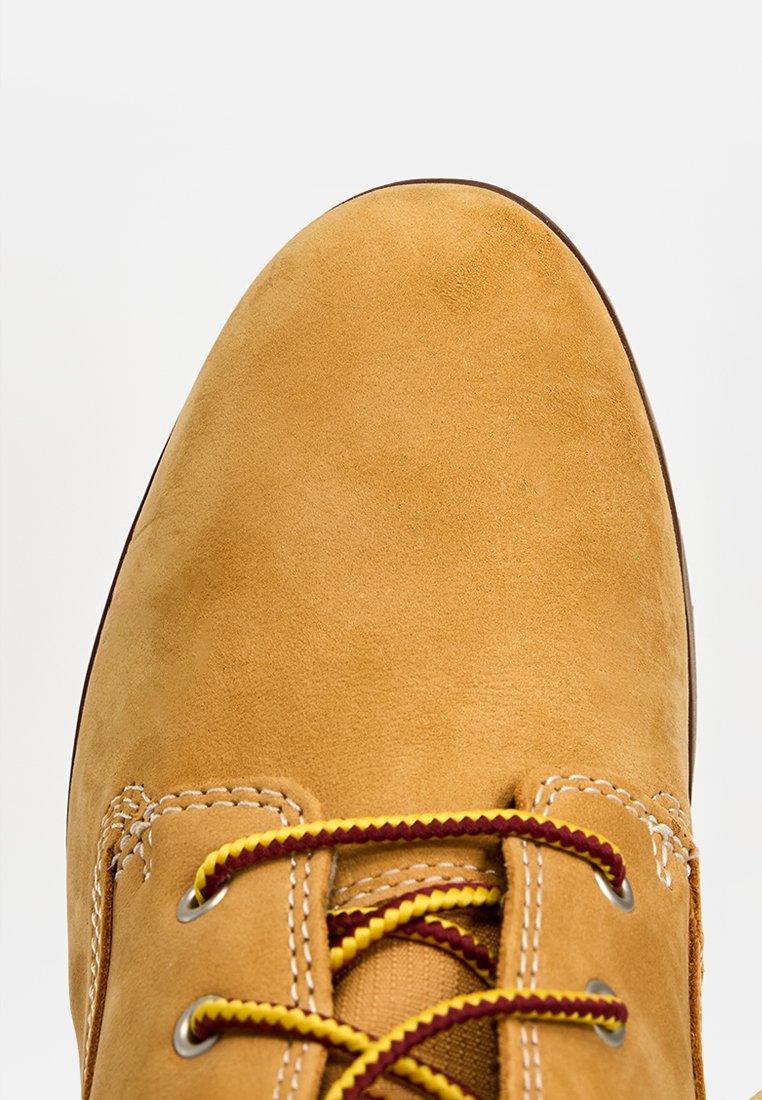 Scarpa scamosciata color beige con punta arrotondata e suola marrone. Presenta lacci gialli e bordeaux e cuciture bianche lungo la parte anteriore.