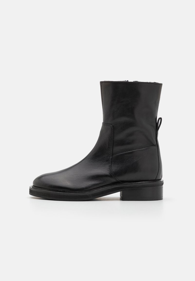 Bottine en cuir noir à texture lisse, embout arrondi et coutures discrètes. Elle possède un talon bloc bas et une languette à l'arrière.