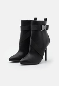 Botines de tacón alto negros en material sintético texturizado, con cintas de tela cruzadas y un acento de hebilla metálica. Diseño de punta afilada.