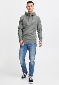 Sudadera gris con botón frontal, capucha ajustable con cordón y puños de punto, combinada con jeans azul desgastados y zapatillas negras.