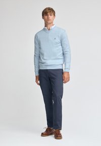 Ljusblå tröja i mjukt material, v-ringning, i kombination med marinblå byxor och bruna snörskor. Svart logotypdetalj på tröjan.