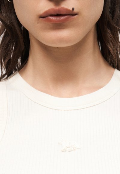 Κοντινό πλάνο από το κάτω μέρος του προσώπου και τον λαιμό ενός ατόμου που φοράει λευκό ριμπ μπλουζάκι με μικρό κεντημένο λευκό ζώο στο στήθος.