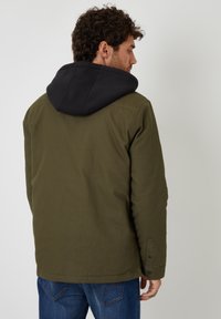 Olivgrön jacka med svart huva, med en slät yta, knäppning vid ärmslut och rak skärning. Stylad för vardagsbruk.