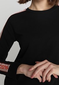 Czarny, długi rękaw bluzki z czerwonymi i białymi paskami na ramionach, z napisem "MAISON" w wyraźnych literach na rękawie. Gładka faktura.