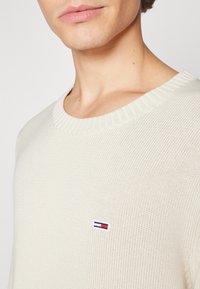 Pleteni pulover u krem boji, s rebrastim ovratnikom i malim logotipom na prsima. Tkanina ima teksturirani, prozračni tkanje.