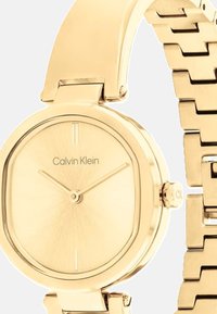 Złoty zegarek Calvin Klein z okrągłą, minimalistyczną tarczą, cienkimi znacznikami godzin i gładką bransoletą. Posiada bezpieczny zamek.