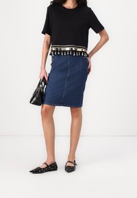 Top negro de manga corta con un dobladillo decorativo de flecos, combinado con una falda lápiz de mezclilla azul con botones laterales y unos zapatos planos negros con acentos de ojales.