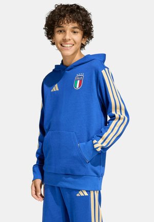 Lächelnder Junge mit lockigem Haar, der einen blauen Italien-Sporthoodie und passende Hose mit goldenen Streifen trägt und mit der Hand in der Tasche steht.