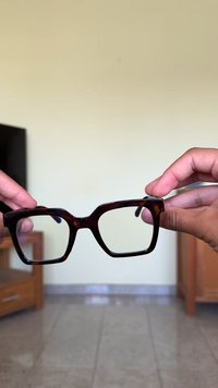 Óculos retangulares de tartaruga segurados por duas mãos. Armação grossa, lentes transparentes e detalhes de textura subtis visíveis.