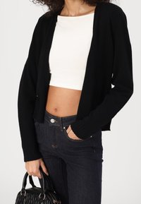 Černý pletený cardigan s krátkým střihem, se žebrovaným lemem a otevřeným předním dílem, v kombinaci s krémovým topem a tmavými džíny.