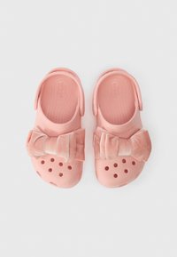 Crocs cor-de-rosa com tiras de calcanhar e grandes laços de tecido macio em cima, exibidos lado a lado sobre um fundo branco.