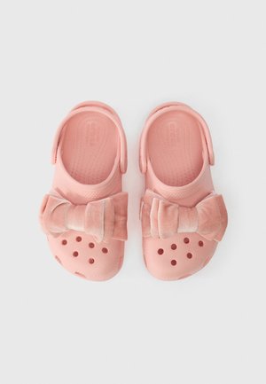 Rožiniai Crocs batai su kulkšnies dirželiais ir didelėmis minkštomis audinio kaspinu, rodomi šalia vienas kito ant balto fono.