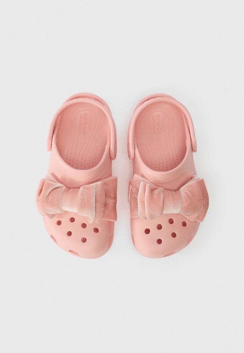 Crocs cor-de-rosa com tiras de calcanhar e grandes laços de tecido macio em cima, exibidos lado a lado sobre um fundo branco.