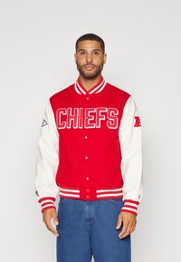 Rde varsity jakna z belimi rokavi, na sprednji strani z besedo "CHIEFS" v krepki beli barvi, zapiranje na gumbe in črtaste rebraste manšete.
